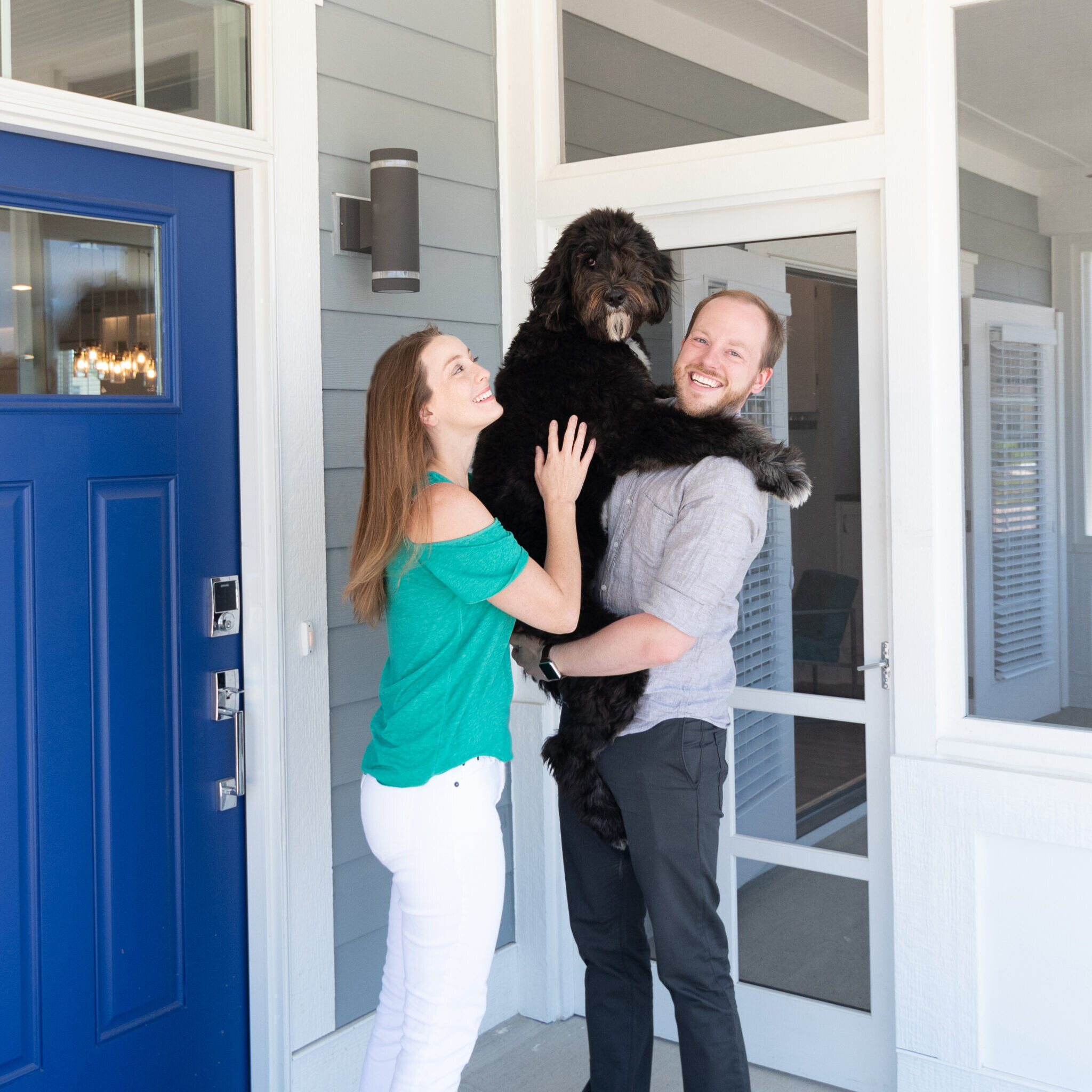 A couple holding a black dog in front of a blue door in their new home.