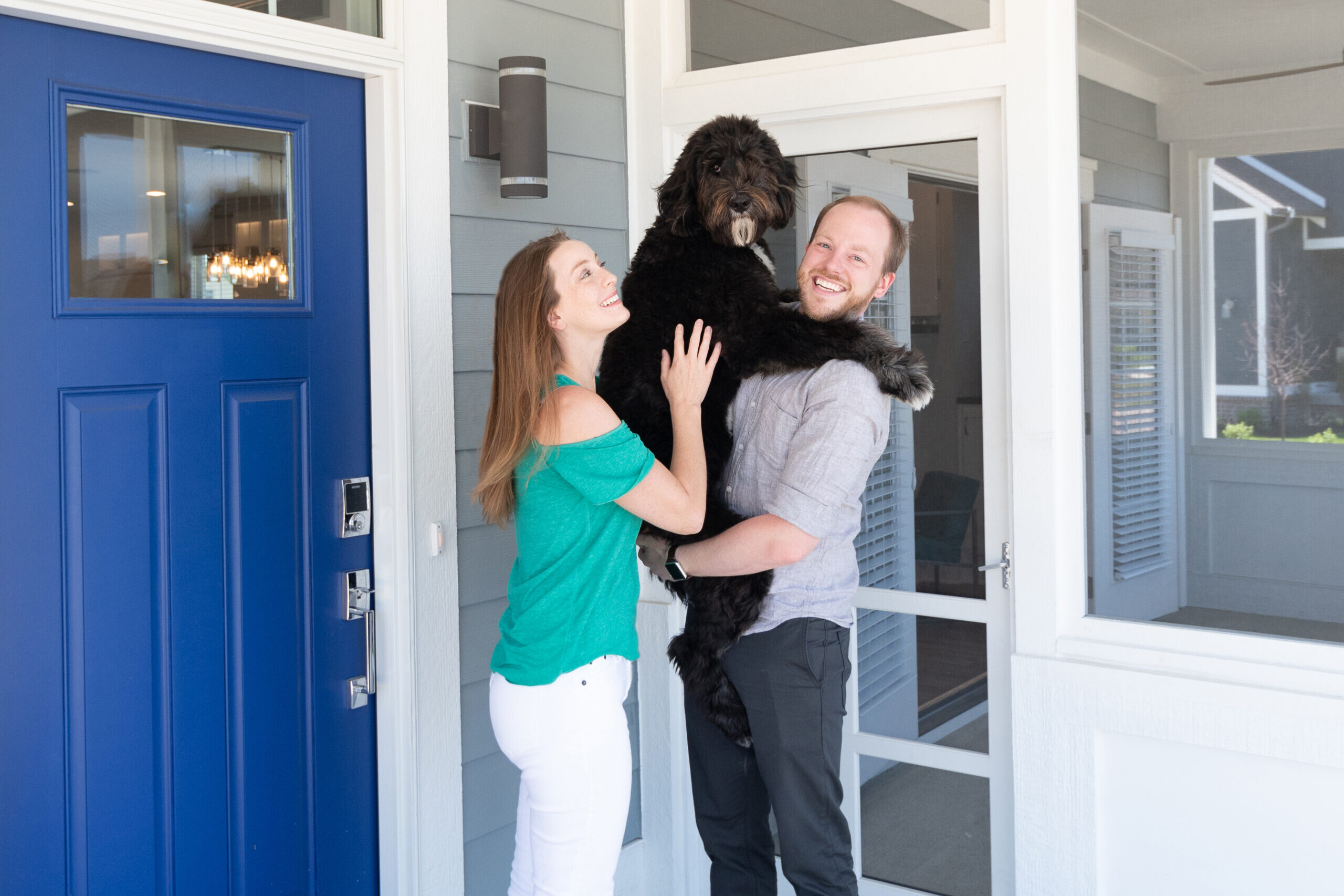 A couple holding a black dog in front of a blue door in their new home.
